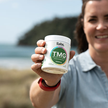 A person holding a container of llabs. TMG Supplement (Betaine), 1000mg, with the ocean and blurred trees in the background, seamlessly incorporating this essential part of their daily routine to enhance overall well-being.