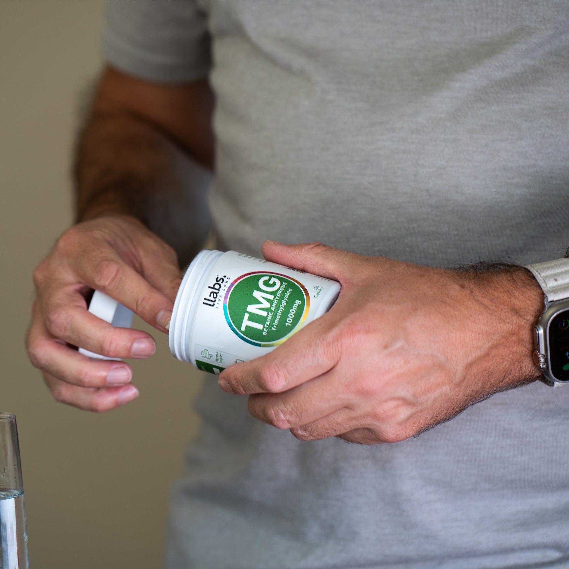 A person in a gray shirt and a smartwatch is holding a container labeled "llabs. TMG Supplement (Betaine)," seamlessly integrating it into their daily routine, with a glass of water partially visible beside them.